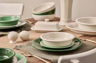 A rustic dining table set with green and cream ceramic dishes, bowls, and plates, along with a butter dish, salt and pepper shakers, gold utensils, and white chairs on a burlap table runner.