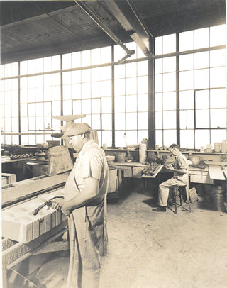 Two men work in a sunlit industrial workshop; one stands at a table shaping material, while the other sits at a bench working with tools. Large windows fill the background.