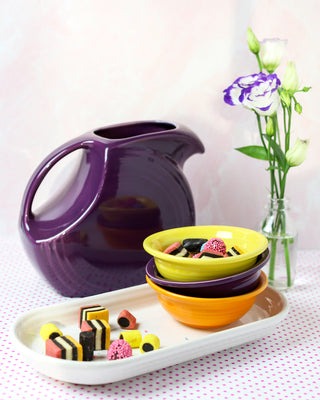A Retired Fiesta 67 OZ Large Disk Pitcher by Fiesta, a small vase with purple and white flowers, two stacked USA-made color glaze bowls, and a white tray with assorted licorice sit on a pink polka dot tablecloth.