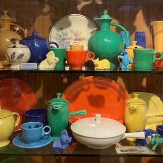 A display shelf filled with colorful vintage dishware, including plates, teapots, cups, and animal figurines in red, yellow, green, blue, orange, and cream, all arranged neatly against a wooden background.