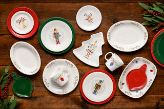 Festive dinnerware set displayed on a wooden table, featuring plates, mugs, and platters with colorful nutcracker and holiday designs, accented with red and green borders and surrounded by greenery and red berries.