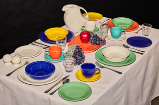 A table set with colorful dishes, cups, and saucers, featuring bright blue, green, yellow, orange, and red. There are glasses, silverware, a white pitcher, salt and pepper shakers, and a bunch of grapes with a red apple.