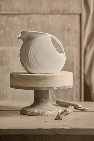 A white ceramic pitcher with circular ridges sits on a pottery wheel covered in clay dust, with sculpting tools lying nearby, against a rustic wooden background.