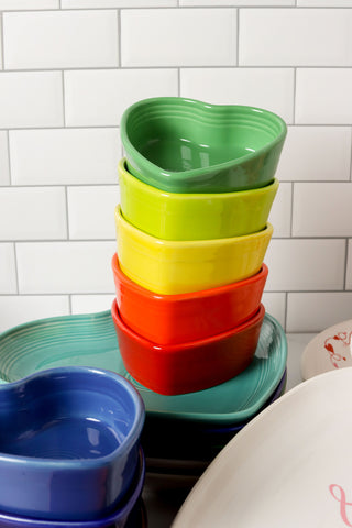 A stack of colorful Fiesta Retired 7 OZ Heart Shaped Ramekins, each 4 1/2 inches in green, yellow, orange, red, and blue ceramic by Fiesta sits on a counter. Made from dishwasher safe vitrified china.