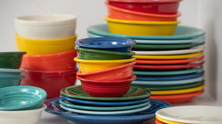 A variety of colorful ceramic bowls and plates are stacked in uneven piles on a white surface, creating a vibrant and cheerful display.