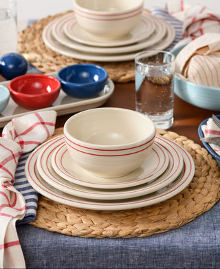 A table setting with Fiesta’s Americana Scarlet Band Classic Bistro 3-Piece Place Setting, Service for 1—cream dishes with red stripes—on a woven placemat with vibrant bowls, blue napkins, bread basket, and blue tablecloth. Made in USA.