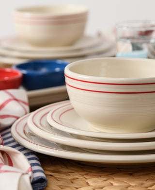 A close-up of Fiesta's Americana Scarlet Band Classic Bistro 3-Piece Place Setting, Service For 1, shows cream-colored plates and bowls with red stripes stacked on a woven placemat, with ramekins, glasses, and striped napkins in the background.