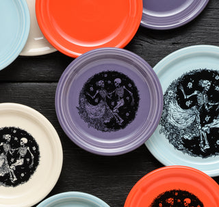 Flat-lay of colorful dinner plates on a black wooden table. Some are plain, others feature a black-ink illustration of two skeletons dancing amidst flowers. Colors include orange, purple, light blue, and cream.
