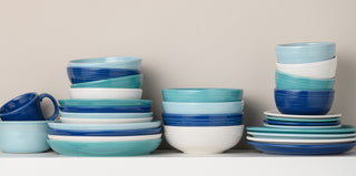 A collection of ceramic bowls, plates, and mugs in shades of blue, turquoise, and white are stacked neatly on a white shelf against a beige wall.