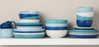 A variety of blue, turquoise, and white bowls, plates, and mugs are stacked neatly on a white shelf against a light beige wall.
