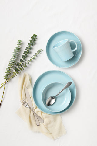 Two light blue plates, a matching cup, and bowl with a spoon sit on a white surface. A fork and knife rest on a folded cream napkin, and a sprig of eucalyptus lies to the left of the dishes.