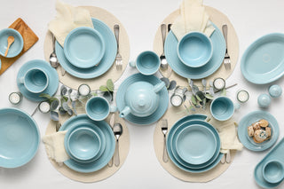 A neatly arranged table setting for four with light blue dishes, bowls, mugs, and a teapot on round placemats. Cream napkins, silver cutlery, candles, and sprigs of greenery decorate the white tablecloth.