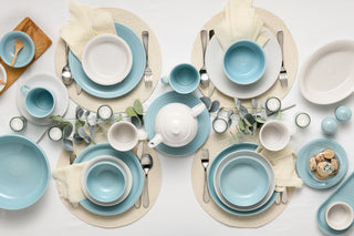 A neatly arranged table setting for four with blue and white plates, bowls, cups, and a teapot on woven placemats. Eucalyptus leaves, tealight candles, and neutral napkins decorate the white tablecloth.