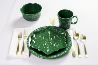A festive green table setting from Fiesta’s Snowflake Collection features the Snowflake Classic Rim 9 Inch Luncheon Plate, gold utensils, a napkin, a bowl, a mug, a candle—all with white snowflake and dot designs on a white tablecloth.