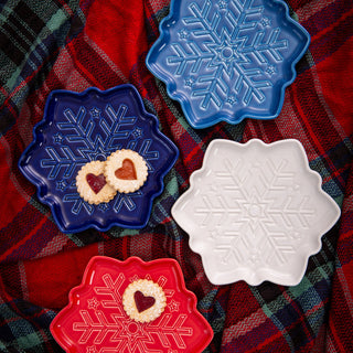 Four Fiesta Snowflake Shaped 9 Inch Plates in blue, light blue, white, and red sit on a red and green plaid blanket. These dishwasher safe plates feature a snowflake design; two hold heart-shaped jam cookies for a festive display.