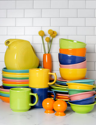 A Retired Fiesta 67 OZ Large Disk Pitcher by Fiesta, along with vibrant ceramic dishes, bowls, and mugs, are neatly stacked on a white marble countertop. In the background is a small orange vase with three yellow flowers.