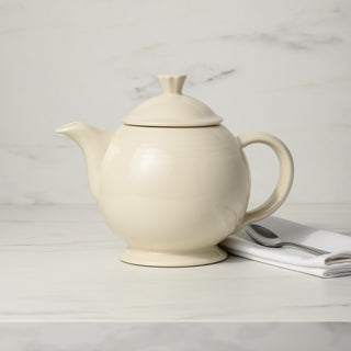 A Retired Fiesta 44 OZ Covered Teapot by Fiesta, in cream-colored lead-safe china, sits on a marble surface near a folded white and gray napkin against a light marble background.