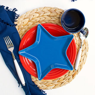 A woven placemat holds a red plate topped with the Fiesta Star Plate, made of fully vitrified china. To the right is a dark blue bowl and spoon; to the left are a fork and napkin for a patriotic-themed hostess gift place setting.