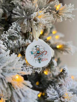 A round Fiesta Nutcracker Sugar Plum Fairy 2026 Ornament hangs on a snowy, decorated Christmas tree with warm yellow lights—a delightful piece of holiday decor.