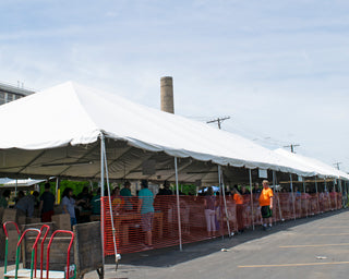 People gather under a large white tent next to orange fencing in a parking lot. Tables and boxes are visible, and a few people wear bright orange shirts. A tall chimney is seen in the background.