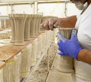 A person wearing a white shirt and blue gloves shapes clay vases on a workbench in a pottery workshop, with several unfinished vases lined up in a row.