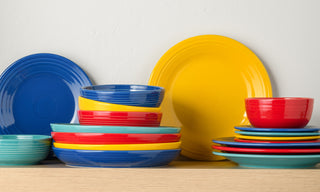 A collection of colorful plates, bowls, and dishes in blue, red, yellow, and teal stacked neatly on a wooden shelf against a plain white wall.
