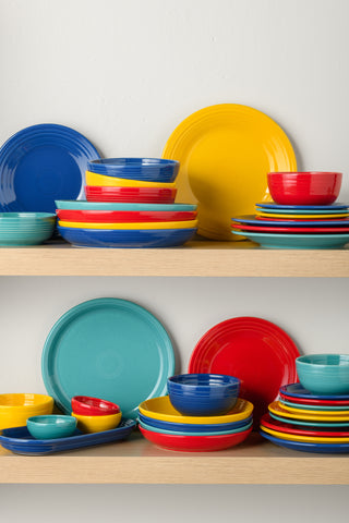 Two wooden shelves hold neatly stacked plates and bowls in bright colors: blue, yellow, red, and turquoise. The vibrant dishware is arranged in an organized and colorful display against a white wall.