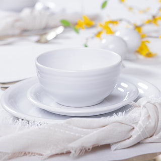 A white ceramic bowl sits on a matching plate and saucer from the Fiesta Bistro Coupe 12-Piece Dinnerware Set, Service for 4, with a white napkin beside it and blurred yellow and white flowers in the background.