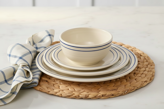 A stack of cream-colored dishes with blue stripes from the Fiesta Americana Lapis Band Classic Bistro 3-Piece Dinnerware Set, Service For 1, rests on a woven placemat with a folded blue-and-white plaid napkin atop a marble surface.