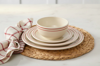 A stack of cream-colored dishes with red stripes, including a bowl from the Fiesta Americana Scarlet Band Classic Bistro 3-Piece Dinnerware Set, Service For 1, sits on a woven placemat beside a folded white napkin with red checks on a marble countertop. Proudly made in the USA.