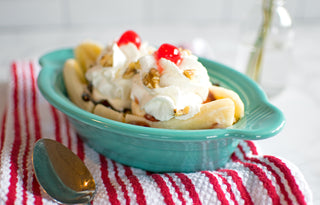 A banana split in a Fiesta 9 1/8 Inch Oval Individual Casserole (13 oz) sits on a red and white striped towel, topped with whipped cream, cherries, and walnuts, next to a spoon.