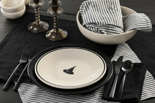 A table setting features black and white plates, one with a witch hat illustration, striped napkins, black cutlery, two candlesticks, and bowls, all arranged on a black placemat and dark wooden table.