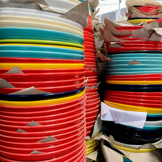 Stacks of colorful ceramic plates in red, yellow, blue, white, and black, separated by sheets of brown paper, arranged closely together in tall piles.