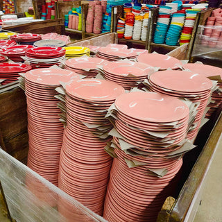 Stacks of pink plates are piled high in wooden crates, surrounded by more stacks of colorful plates in red, yellow, blue, and white, inside what appears to be a warehouse or store.