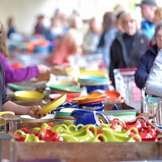 People browse and select colorful plates and cups from bins at a busy pottery sale, with shoppers in the background and stacks of vibrant dinnerware in the foreground.
