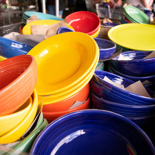 Stacks of colorful ceramic bowls and plates in yellow, orange, blue, red, and green, arranged closely together on a table, with paper napkins placed between some of the dishes.
