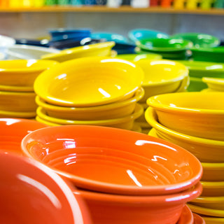 Stacks of brightly colored ceramic bowls in orange, yellow, green, and blue, displayed on a table with more colorful dishes blurred in the background.