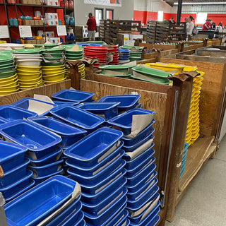Stacks of colorful plastic trays—blue, yellow, green, and red—are organized on wooden shelves inside a store or warehouse. People are visible in the background near shelves and product displays.