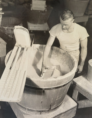 A man in a white shirt stands beside a large wooden vat, stirring a creamy liquid with a tool; another container and equipment are nearby in what appears to be a factory or production setting.