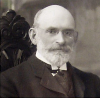 An older man with a bald head, white beard, and glasses, wearing a suit and sitting on a carved wooden chair, poses for a formal portrait in black and white.