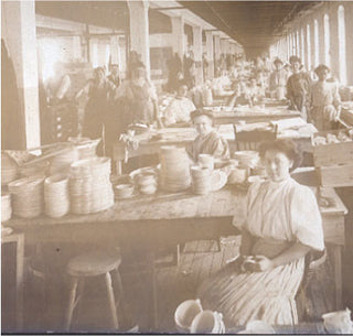 A sepia-toned photo shows women working at long tables in a factory lined with stacks of bowls. The workspace is crowded and lit by tall windows on one side, with workers busy handling ceramics.