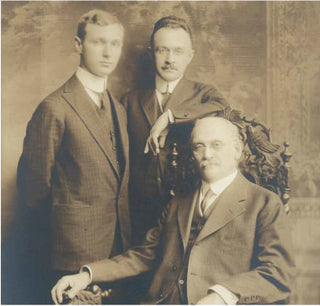 Three men in formal suits pose for a vintage sepia-toned portrait. Two stand behind, one with a hand on the shoulder of the seated older man, who rests in an ornate wooden chair. All have serious expressions.