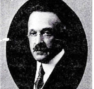 Black and white oval portrait of a middle-aged man with a mustache, wearing a suit and tie, looking slightly to the side. The background appears dark and grainy.