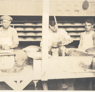 Three men work with clay at a pottery workshop. Two wear aprons, and one is in a tank top. Shelves of pottery pieces are visible behind them. The setting appears old-fashioned and industrial.