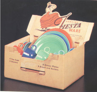 A cardboard box labeled Genuine Fiesta Ware holds colorful ceramic dishes, including plates, cups, and saucers. The box features an illustration of a dancing woman in a flowing dress on the lid.