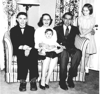 A black-and-white photo of a family of five posing on a striped sofa. Two adults sit in the middle, holding a baby, with a boy seated on the left and a girl standing on the right, all dressed formally.