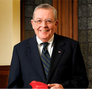 An older man wearing glasses, a dark suit, striped tie, and an American flag pin smiles at the camera while standing indoors in front of wood and glass paneling.