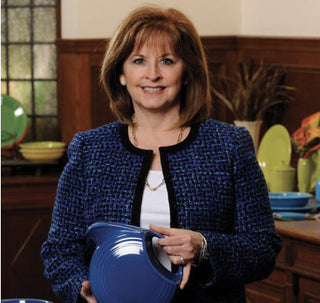 A woman with shoulder-length brown hair, wearing a blue textured jacket, stands indoors holding a blue ceramic dish. Colorful dishes and a window with a wooden frame are visible in the background.