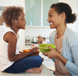 A woman and a young girl sit facing each other, smiling and holding bowls of food. The girl sits on a kitchen counter while the woman holds a green bowl, both enjoying a happy moment together.