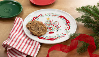 A chocolate chip cookie rests on the Fiesta Holly Berry Christmas Tree 12 Inch Round Baking and Serving Platter, alongside a red-and-white striped napkin, green and red holiday dinnerware, and pine branches on a wooden table.